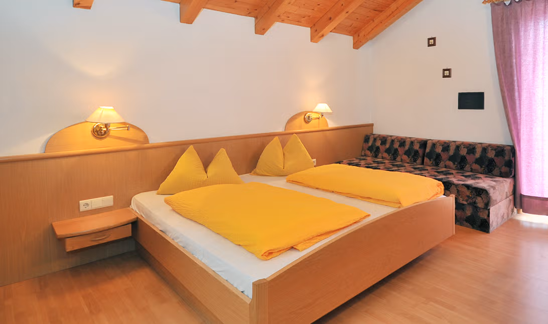 Gemütliches Doppelzimmer des Brunnerhofs in der Val Pusteria, mit Naturholzbetten, gelber Bettwäsche und Sofa.