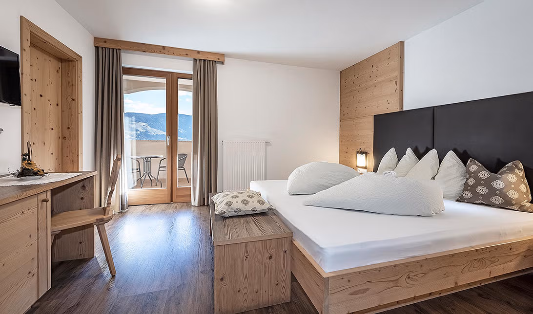 Doppelzimmer aus Naturholz mit Bett, Schreibtisch und Balkon mit Blick auf die verschneiten Berge.