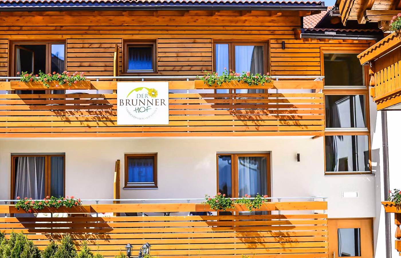 Holzfassade des Brunnerhofs mit blühenden Balkonen und dem Logo, das zu einem Bauernhofurlaub im Pustertal einlädt.