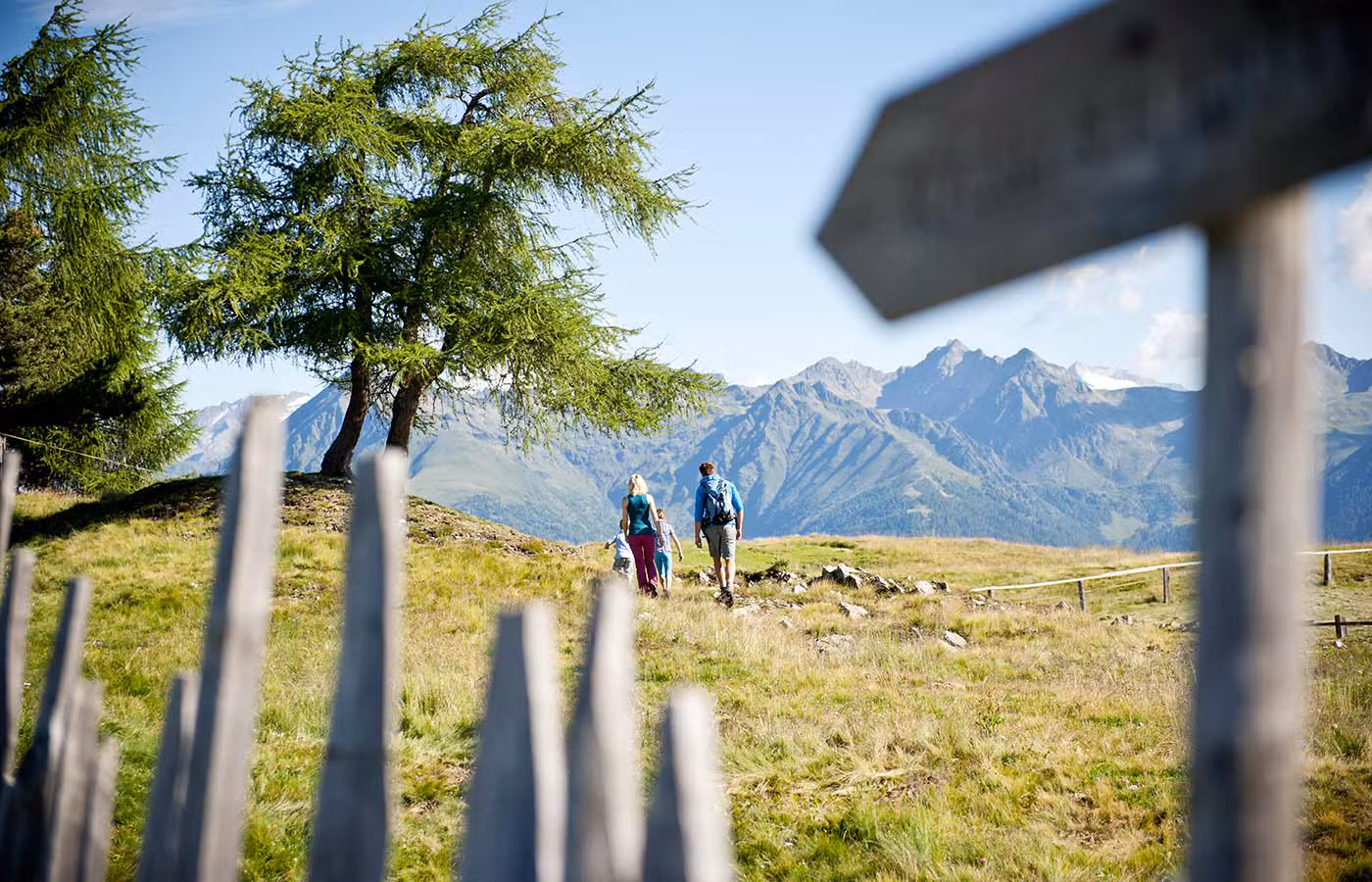 Famiglia in escursione su un sentiero erboso in Val Pusteria, con montagne maestose e un cartello in primo piano.