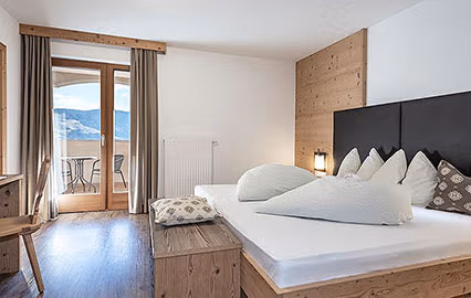 Camera Eichenkrone dell'agriturismo Brunnerhof, con letto matrimoniale in legno naturale, cuscini bianchi e balcone con vista sulle montagne della Val Pusteria.