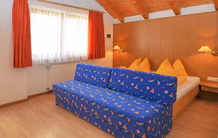 Accogliente camera Eichenkrone con divano letto blu, letto matrimoniale e pareti in legno naturale, ideale per vacanze in famiglia in Val Pusteria.