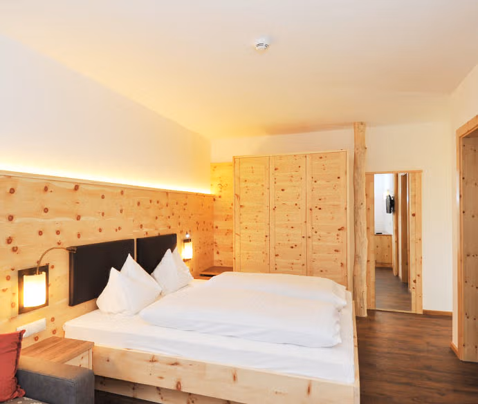 Camera matrimoniale Zirmspitz con arredi in legno naturale di abete rosso e cirmolo, letto con lenzuola bianche, luci calde e armadio a muro, tipica dell'agriturismo in Val Pusteria.
