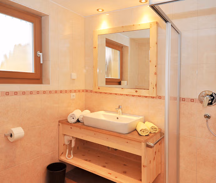 Bagno moderno e luminoso della camera Zirmspitz con lavabo in ceramica, specchio con cornice in legno naturale e doccia.