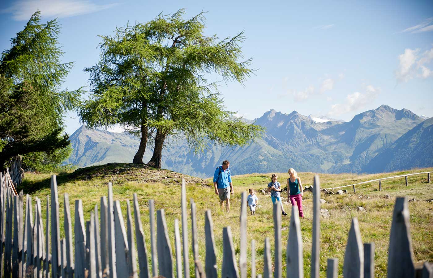 famigliola che passeggia in un prato alpino della Val Pusteria con uno steccato in primo piano e i monti sullo sfondo