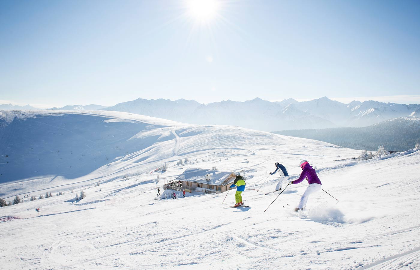 Skifahrer fahren auf einer Piste ins Tal: Berge und glasklarer blauer Himmel im Hintergrund