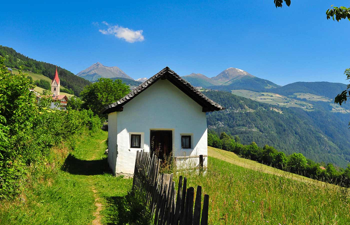 Una casetta solitaria in un mezzo a un prato di montagna con una chiesetta sullo sfondo